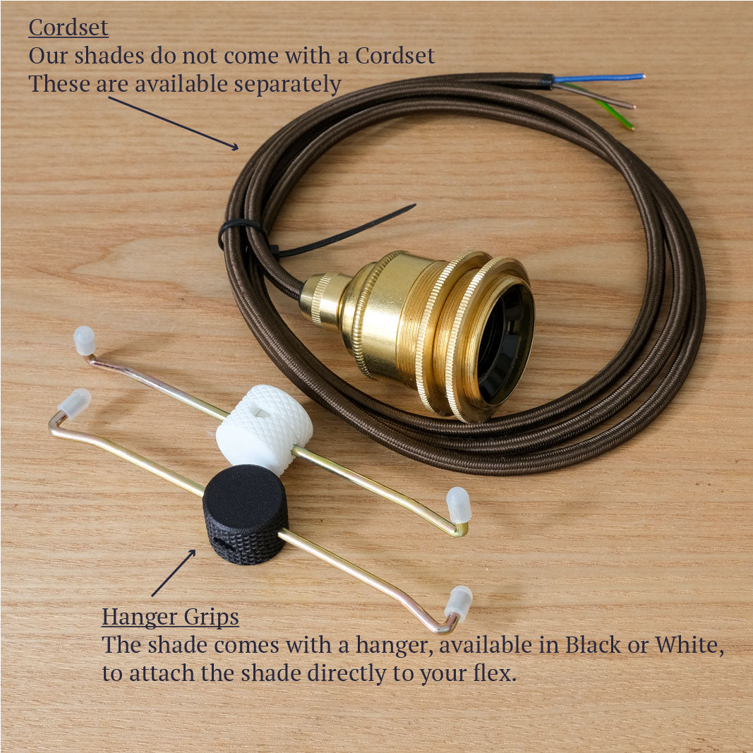 A cordset light fixture component set laid out on a light wood background, including a coiled brown cord with a brass socket, two metal hanger grips, a white plastic cap, and a black cylindrical piece. Text labels identify the cordset and hanger grips and note that shades are sold separately.